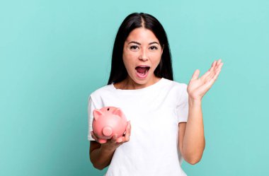 hispanic pretty woman feeling happy and astonished at something unbelievable with a piggy bank