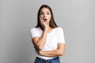 Güzel, genç bir kadın şok ve inançsızlıkla ağzı açık, elleri yanağında ve kolları çaprazlanmış, sersemlemiş ve şaşırmış hissediyor.