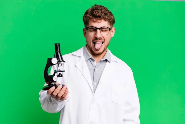 陽気で反抗的な態度を持つ若い大人の白人男性冗談と舌を突き出す 顕微鏡の概念を持つ科学研究室の学生は — ストック写真