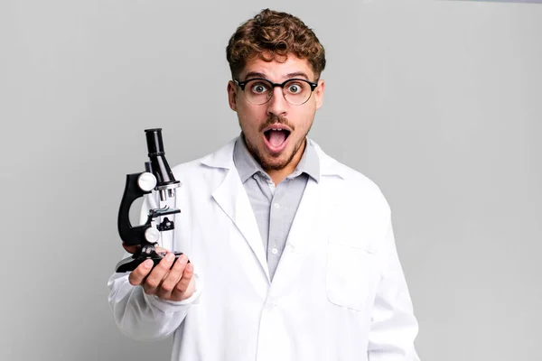 年轻的高加索男子看上去非常震惊或惊讶 带有显微镜概念的科学实验室学生 — 图库照片