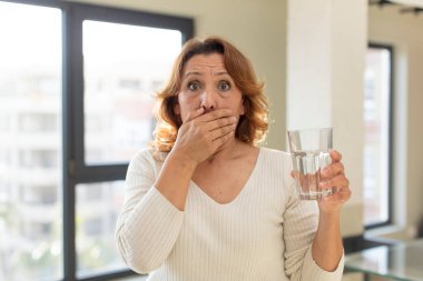 Orta yaşlı, güzel bir kadın ağzını eliyle kapatıyor ve şok olmuş ya da şaşırmış bir ifade takınıyor. su bardağı