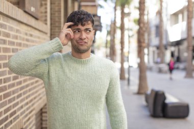 Genç yakışıklı adam şaşkın ve şaşkın hissediyor, başını kaşıyor ve yana bakıyor.