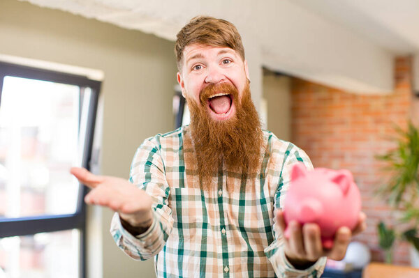 red hair man smiling happily and offering or showing a concept with a piggy bank
