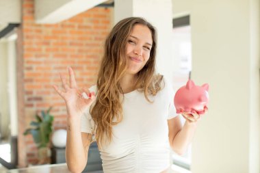 Güzel bir kadın kendini mutlu hissediyor, iyi bir jestle onaylıyor. Domuz kumbarası konsepti