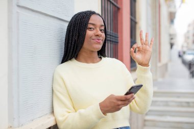 Siyah afro kadın kendini mutlu hissediyor, iyi bir jestle onaylıyor. akıllı telefon konseptini kullanarak