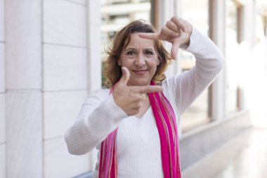 Orta yaşlı bir kadın mutlu, arkadaş canlısı ve pozitif hissediyor, gülümsüyor ve elleriyle bir portre veya fotoğraf çerçevesi yapıyor.