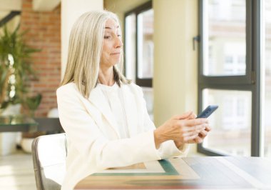 Güzel, kıdemli bir kadın gülümsüyor ve mutlu, kendinden emin bir ifadeyle akıllı bir telefonla bakıyor.