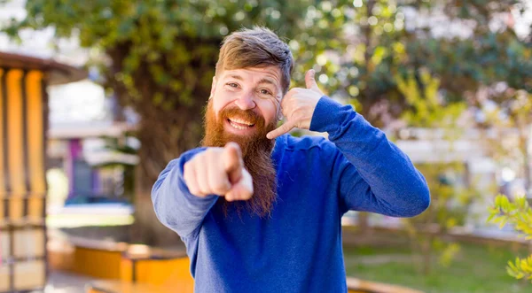 stock image red hair bearded man smiling cheerfully and pointing to camera while making a call you later gesture, talking on phone