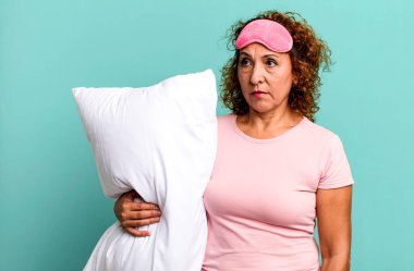 Güzel orta yaşlı bir kadın üzgün, üzgün ya da kızgın ve pijama geceliği ve yastıkla yan tarafa bakıyor.