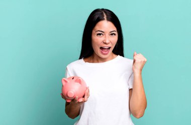 hispanic pretty woman feeling shocked,laughing and celebrating success with a piggy bank