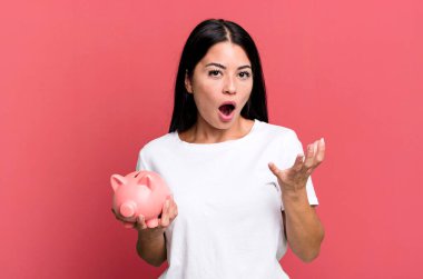 hispanic pretty woman amazed, shocked and astonished with an unbelievable surprise with a piggy bank