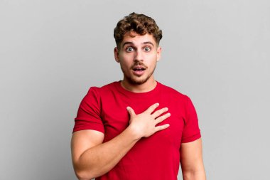 young adult caucasian man feeling shocked, astonished and surprised, with hand on chest and open mouth, saying who, me?