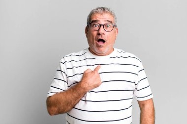Orta yaşlı bir adam şok olmuş ve ağzı açık bir şekilde kendini işaret ederek şaşırmış görünüyor.
