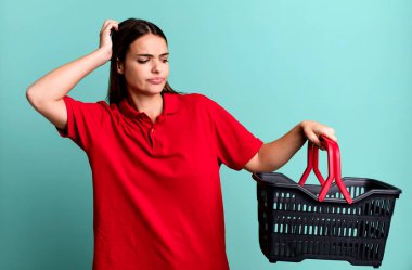 young pretty woman smiling happily and daydreaming or doubting. empty shopping basket concept