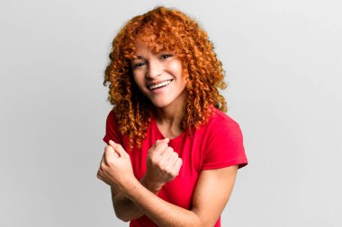 kızıl saçlı güzel kadın neşeli bir şekilde gülümsüyor ve kutluyor, yumruklarını sıkıp kollarını kavuşturuyor, mutlu ve pozitif hissediyor.