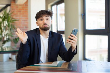 Genç yakışıklı adam omuz silkiyor, kafası karışık ve kararsız. akıllı telefon kavramı