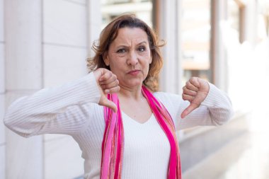 Orta yaşlı bir kadın üzgün, hayal kırıklığına uğramış ya da kızgın görünüyor. Anlaşmazlıkta baş parmağını gösteriyor. Hayal kırıklığına uğramış hissediyor.