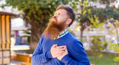 Kızıl sakallı adam üzgün, yaralı ve kalbi kırık görünüyor, iki elini de kalbine yakın tutuyor, ağlıyor ve depresif hissediyor.