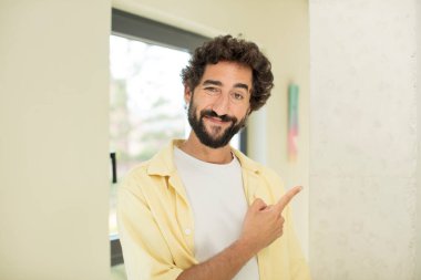 Genç, çılgın sakallı, neşeli bir şekilde gülümseyen, mutlu hisseden, yan ve yukarı bakan, fotokopi uzayındaki nesneleri gösteren bir adam.