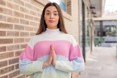 pretty young adult woman feeling worried, hopeful and religious, praying faithfully with palms pressed, begging forgiveness