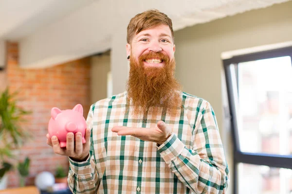 红头发的男人兴高采烈地笑了笑 感到快乐 并和小猪银行展示了一个概念 — 图库照片
