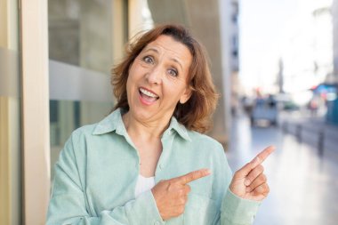 Orta yaşlı kadın neşeyle gülümsüyor ve yan taraftaki fotokopi alanına işaret ediyor, bir nesneyi gösteriyor veya reklamını yapıyor.