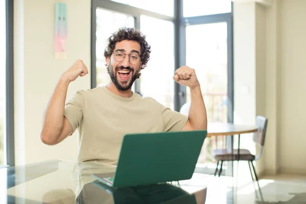 stock image young adult bearded man with a laptop feeling happy, satisfied and powerful, flexing fit and muscular biceps, looking strong after the gym