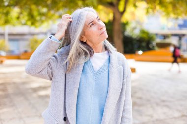Yaşlı, emekli, güzel, beyaz saçlı, kafası karışmış, başını kaşıyan ve yan tarafa bakan bir kadın.