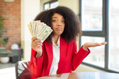 Oldukça afro siyah bir kadın omuz silkiyor, kafası karışık ve kararsız hissediyor. Dolar banknotları konsepti