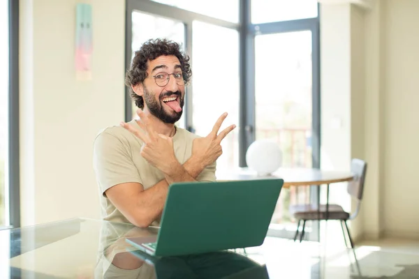 一个留着胡子的年轻人 带着笔记本电脑 面带微笑 看上去快乐 用双手表示胜利或和平 — 图库照片