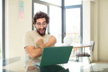 Genç, sakallı, dizüstü bilgisayarlı, neşeli bir şekilde gülümseyen ve kollarını kavuşturup kollarını kavuşturan mutlu ve pozitif hisseden bir adam.