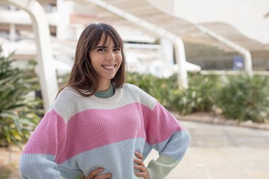 Güzel genç bir kadın mutlu bir şekilde gülümsüyor. Elleri kalçasında ve kendinden emin, gururlu ve arkadaş canlısı bir tavırla.