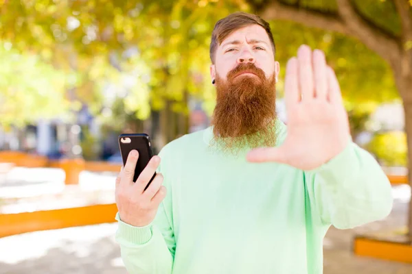 年轻的留着胡子的男人看上去很严肃 用智能手机摆出张开手掌的姿势 — 图库照片