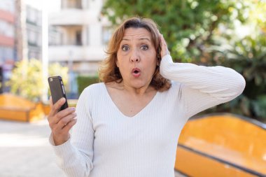 Orta yaşlı güzel bir kadın son derece şaşkın ve şaşırmış hissediyor. Akıllı telefonunu kullanarak.