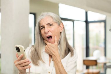 Ağzı ve gözleri sonuna kadar açık ve eli çenesinde güzel bir son sınıf öğrencisi. akıllı telefon kavramı