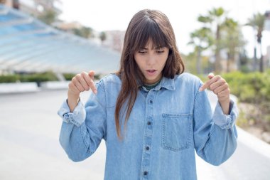 Ağzı açık, iki eliyle aşağıya doğru bakan güzel genç bir kadın. Şok olmuş, şaşırmış ve şaşırmış görünüyor.