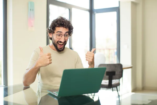一个留着胡子的年轻人 带着笔记本电脑 笑容满面 看上去很快乐 无忧无虑 两根大拇指都挺直了 — 图库照片