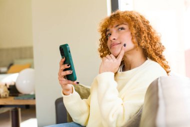 Genç kızıl saçlı, Latin güzel bir kadın evde akıllı telefon kullanıyor.