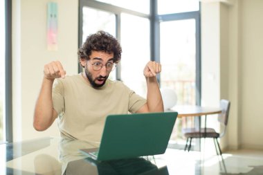 Diz üstü bilgisayarı olan sakallı genç bir adam iki eliyle aşağıya bakıyor, şaşkın, şaşkın ve şaşırmış görünüyor.