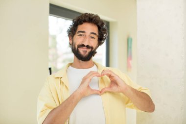 Genç çılgın sakallı adam gülümsüyor ve mutlu, sevimli, romantik ve aşık, iki eliyle kalbi şekillendiriyor.