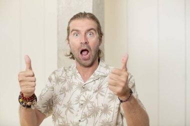 Genç, sarışın, yetişkin bir adam neşeyle gülümsüyor ve mutlu görünüyor. Kaygısız ve pozitif hissediyor. İki başparmağı da havada.