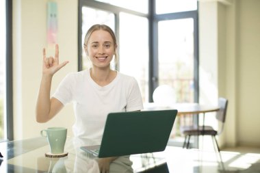 Dizüstü bilgisayarı olan beyaz bir kadın mutlu, eğlenceli, kendinden emin, pozitif ve isyankar, elle taş ya da ağır metal işareti yapıyor.