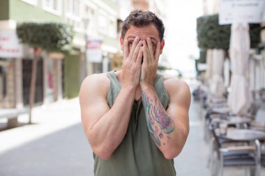 Yakışıklı adam elleriyle yüzünü kapatıyor, parmaklarının arasından şaşkın bir ifadeyle yan tarafa bakıyor.