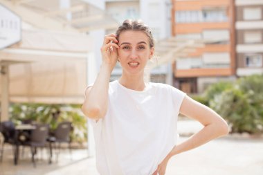 Güzel bir kadın şok ve inançsızlık içinde, elleri yanağında ve kolları çaprazlanmış, sersemlemiş ve şaşırmış hissediyor.