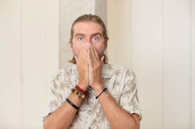 Genç sarışın yetişkin adam mutlu, neşeli, şanslı ve şaşkın görünüyor. İki eliyle ağzını kapatıyor.