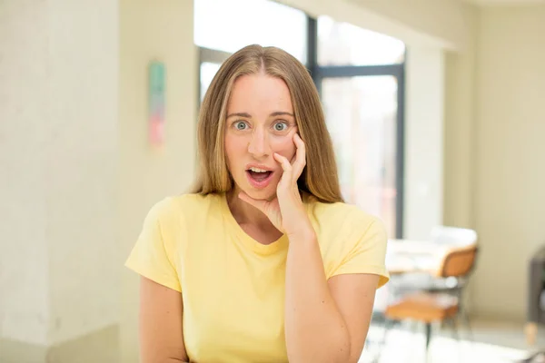 stock image pretty blond woman open-mouthed in shock and disbelief, with hand on cheek and arm crossed, feeling stupefied and amazed