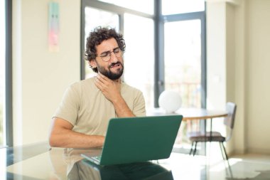 Genç, sakallı, dizüstü bilgisayarı olan, boğazı ağrıyan ve grip belirtileri gösteren ağzı kapalı öksüren bir adam.