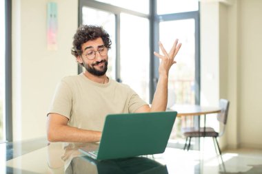 Genç, sakallı, laptoplu bir adam sabahın erken saatlerinde uykulu, yorgun ve sıkılmış görünüyor.