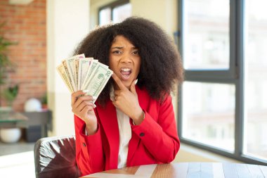 Ağzı ve gözleri açık, elleri çenesinde güzel afro bir zenci kadın. Dolar banknotları konsepti