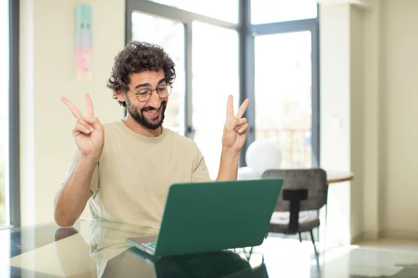 一个留着胡子的年轻人 带着笔记本电脑 面带微笑 看上去快乐 用双手表示胜利或和平 — 图库照片